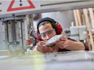 homme avec casque anti bruit près d'une machine industrielle qui tient et regarde une pièce