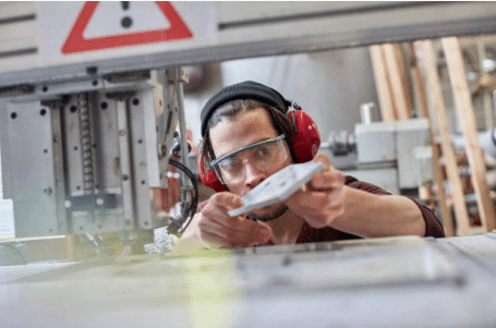 homme avec casque anti bruit près d'une machine industrielle qui tient et regarde une pièce